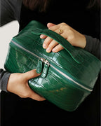 Woman holding a green layflat leather vanity bag with a zippered closure and handle.