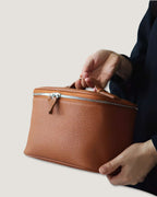 Medium leather vanity bag being held, showcasing its textured full grain leather and metal zip-around closure.